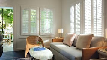 A living room with a sofa, a wicker chair, a glass coffee table, and a lamp. There are large windows with shutters and a view of the outdoors.