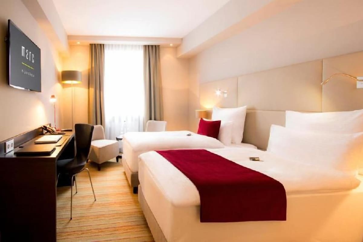 Une chambre d’hôtel avec deux lits, un bureau, une chaise, une télévision et une fenêtre avec des rideaux.