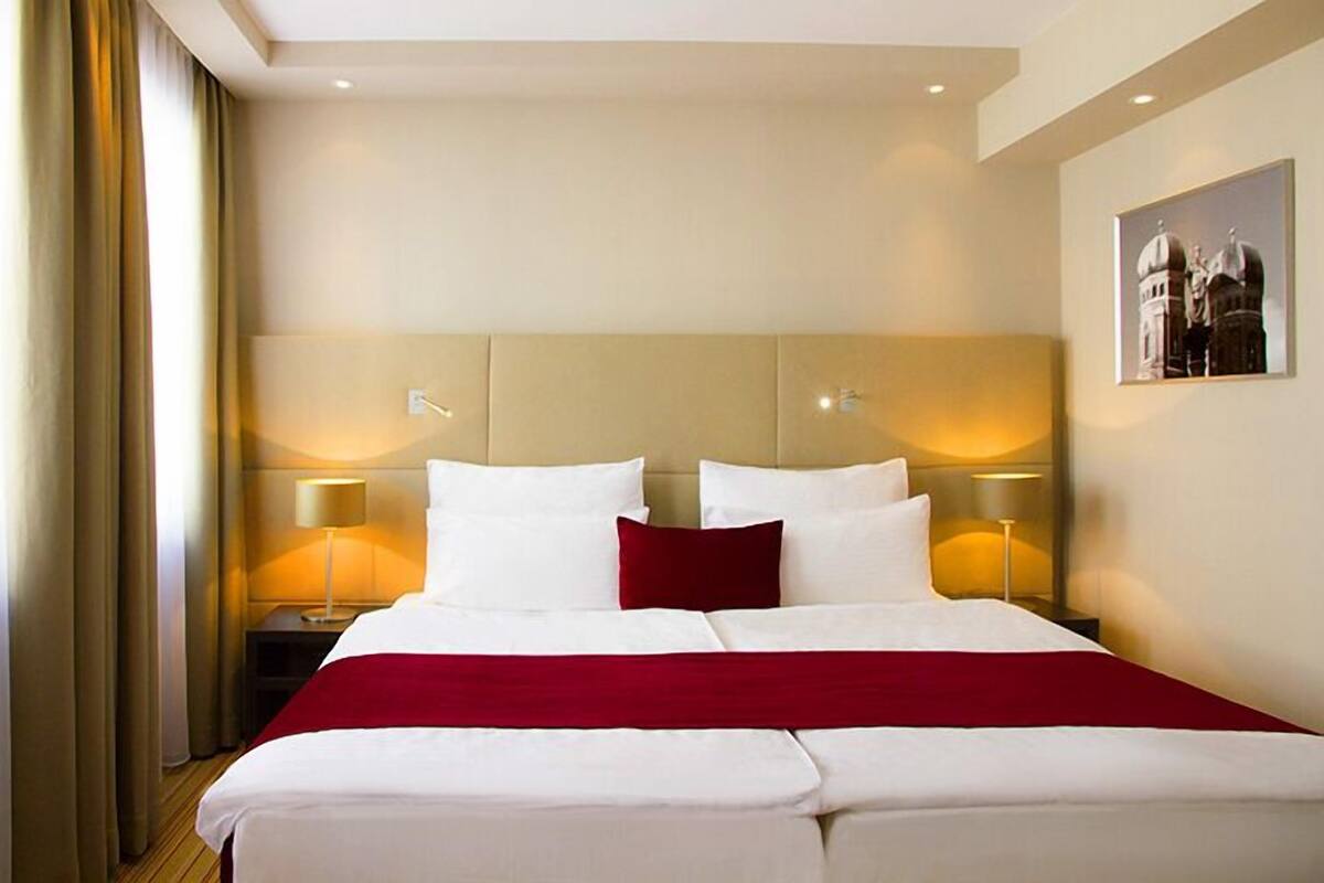 A hotel room with a large bed, two bedside lamps, a red throw blanket, and a framed picture on the wall.
