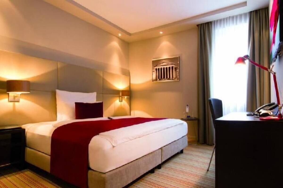 A hotel room with a bed, bedside lamps, a desk, and a window with curtains.
