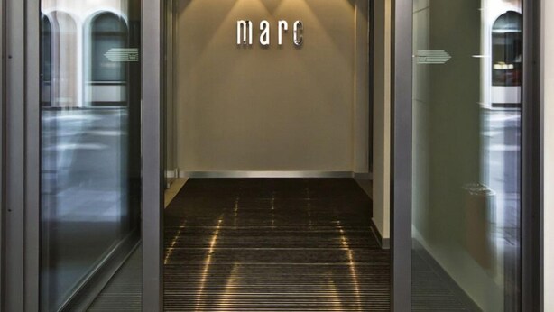 A modern elevator entrance with a glass door, a metallic handrail, and a wall-mounted light fixture. - marc münchen - Adults Only (Munich)