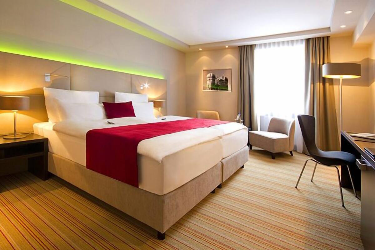 A hotel room with a large bed, a desk, a chair, and a desk lamp.