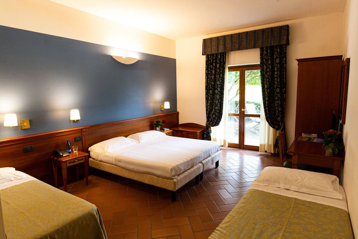 Habitación de hotel con dos camas individuales, cabecero de madera, mesita de noche con lámpara, una pequeña mesa con una planta y vista al exterior a través de una puerta con cortinas.