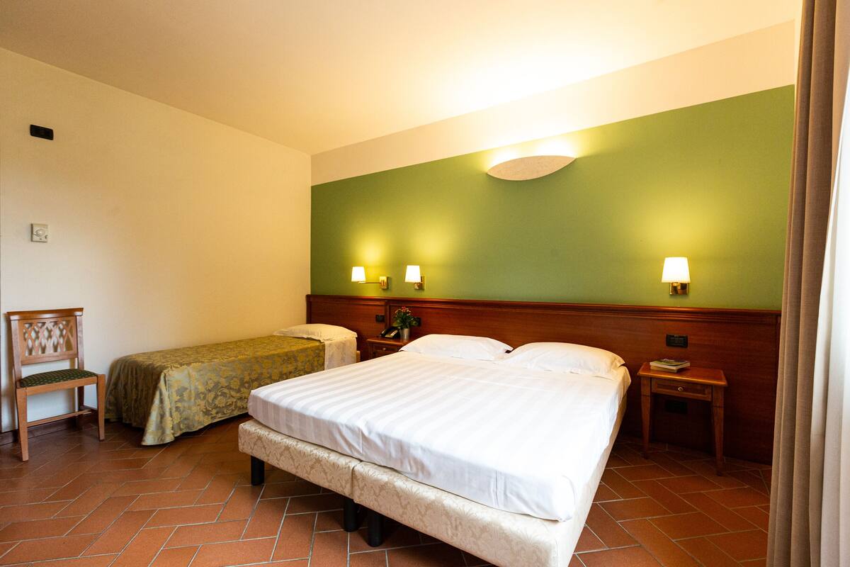 Habitación de hotel con dos camas individuales, una mesita de noche de madera, una silla y una pared verde decorativa.