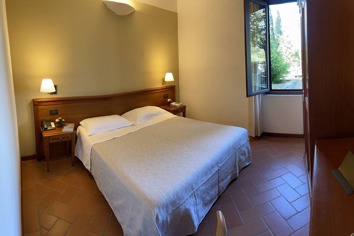 Habitación de hotel con cama, lámparas de noche, una mesita con teléfono y un libro, ventana con vistas a zonas verdes y televisor montado en la pared.