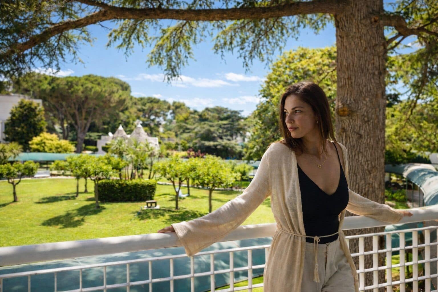 A woman standing on a balcony with a view of a park and buildings in the background.