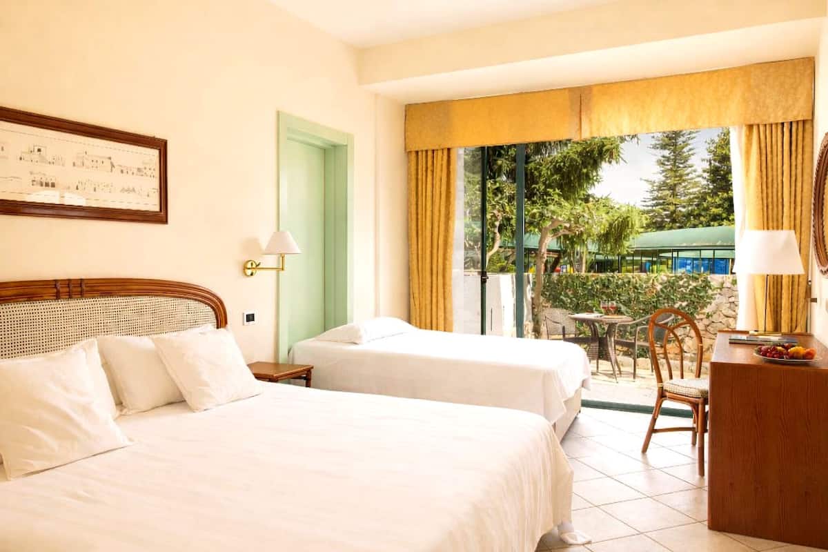 Une chambre d’hôtel avec deux lits, un balcon donnant sur un espace verdoyant et un bâtiment, un bureau avec une chaise et un tableau encadré au mur.