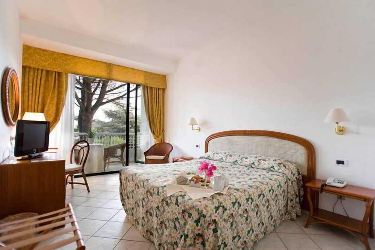 A hotel room with a bed, a desk with a computer, a chair, a small table with a phone, and a window with curtains.