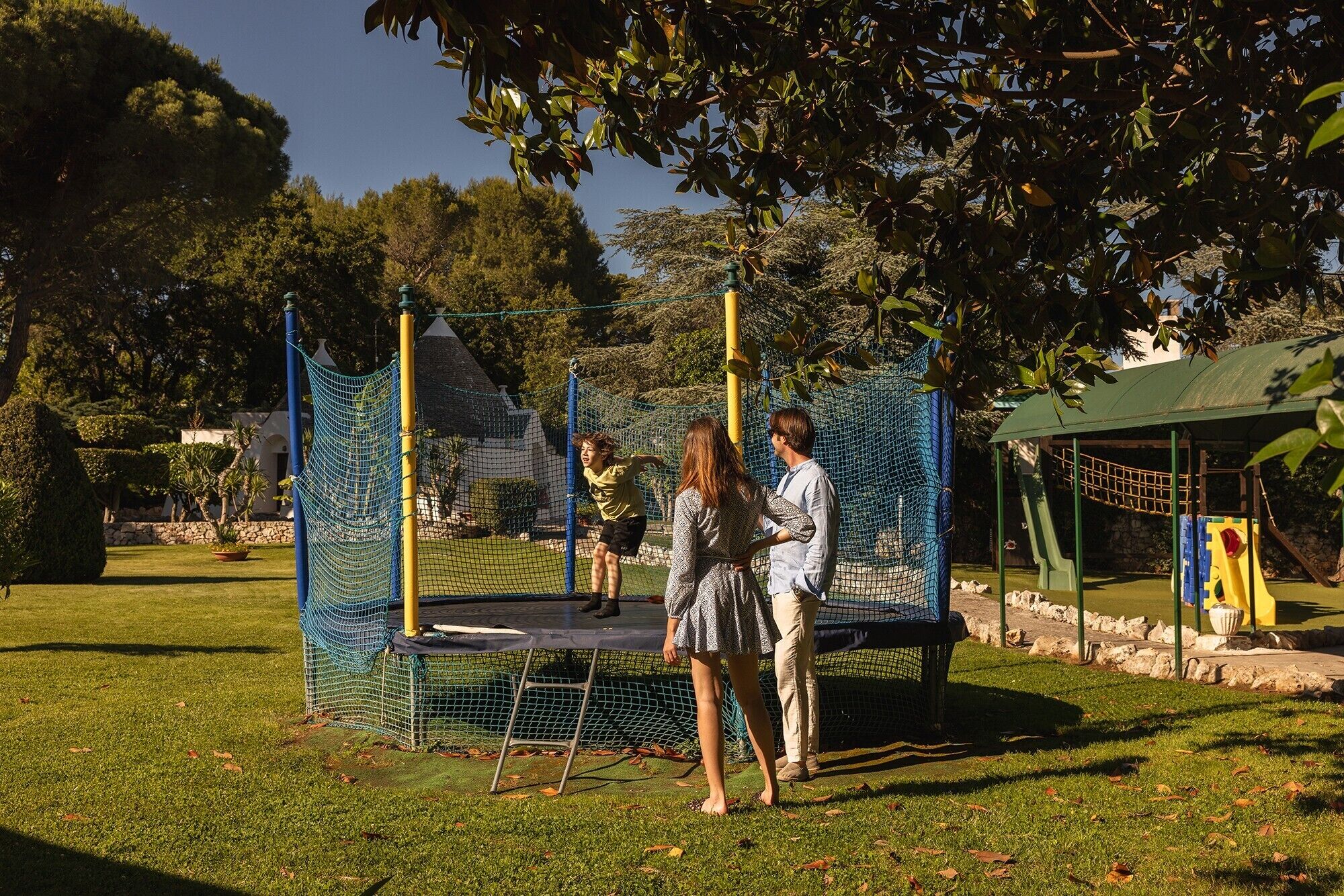 A playground with a trampoline and netted climbing structures, surrounded by grass and trees.