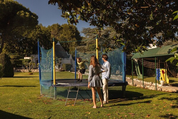 A playground with a trampoline and netted climbing structures, surrounded by grass and trees.