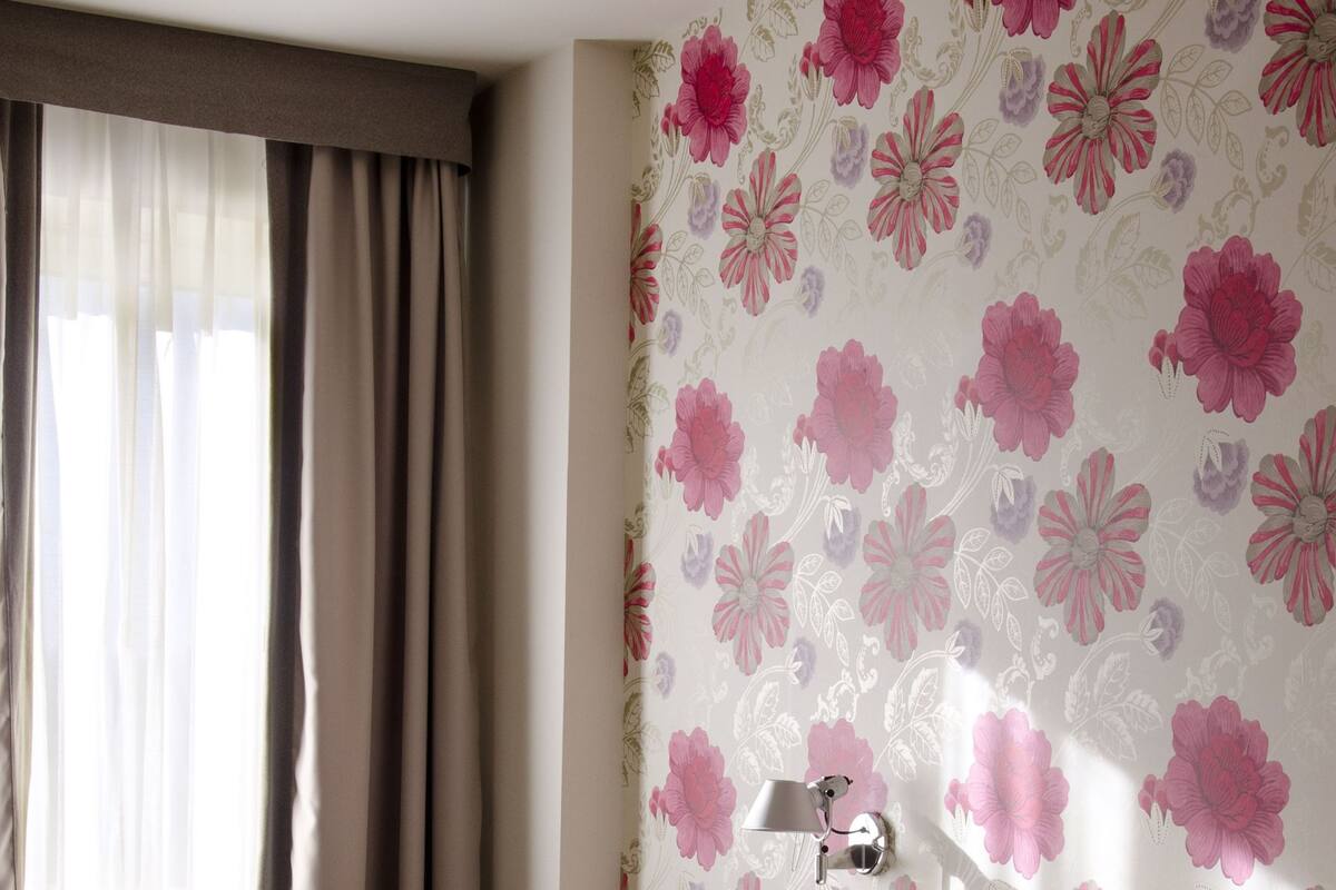Una camera d'albergo con un letto, un comodino, una lampada e carta da parati con motivi floreali.