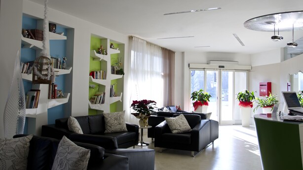 A modern living room with two black sofas, a coffee table, and a bookshelf. There is a large window with curtains, a door leading outside, and a small table with a vase of flowers. - Hotel Key (Vicenza)