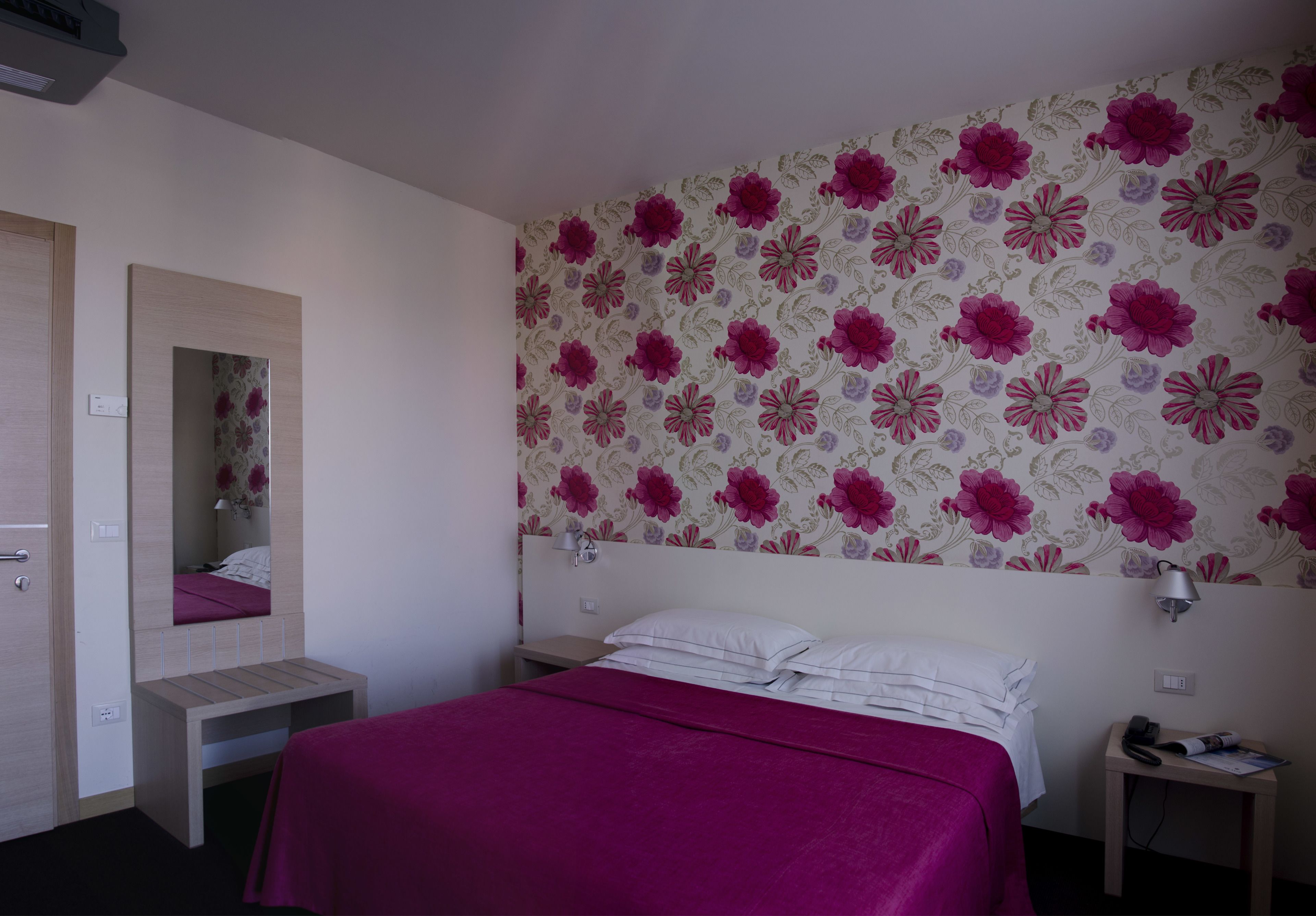 Un dormitorio con cama, mesita de noche, espejo y papel tapiz floral en la pared.