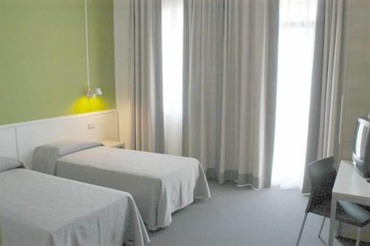 Camera d'albergo con due letti singoli, una scrivania con sedia, una televisione e una finestra con tende.