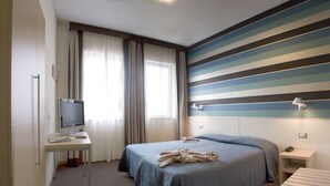 A hotel room with a bed, a desk with a television, a bedside table with a lamp, and a striped wall. - Hotel Key (Vicenza)