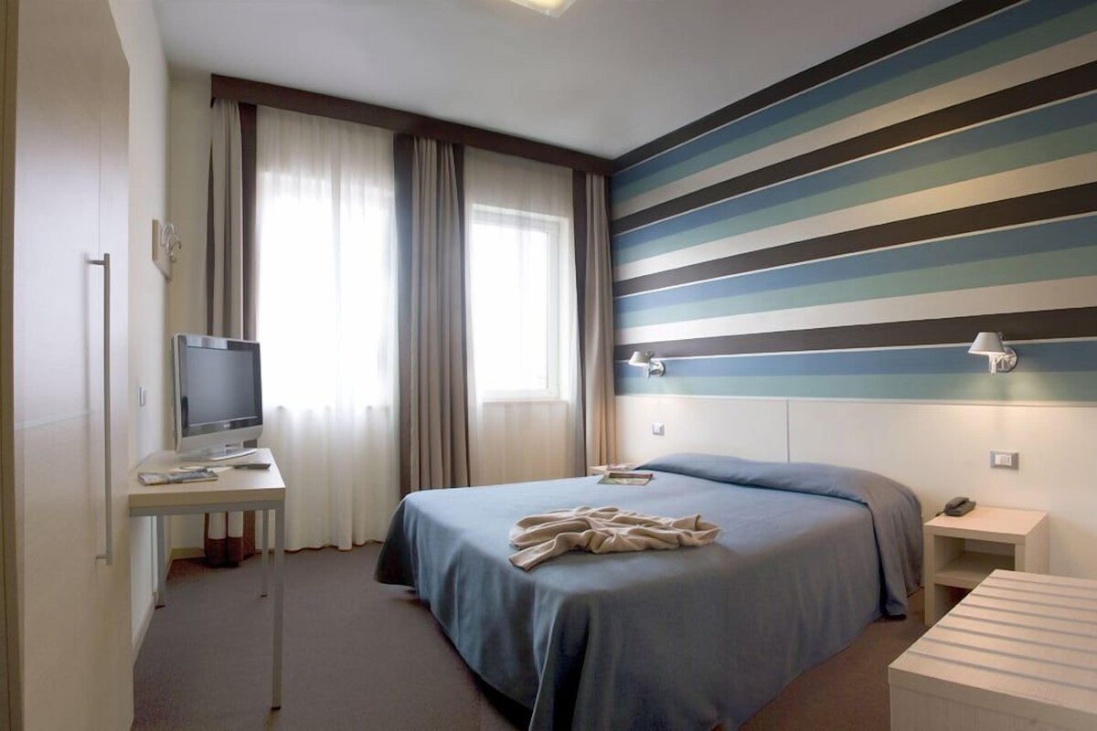 A hotel room with a bed, a desk with a television, a bedside table, and a striped wall.