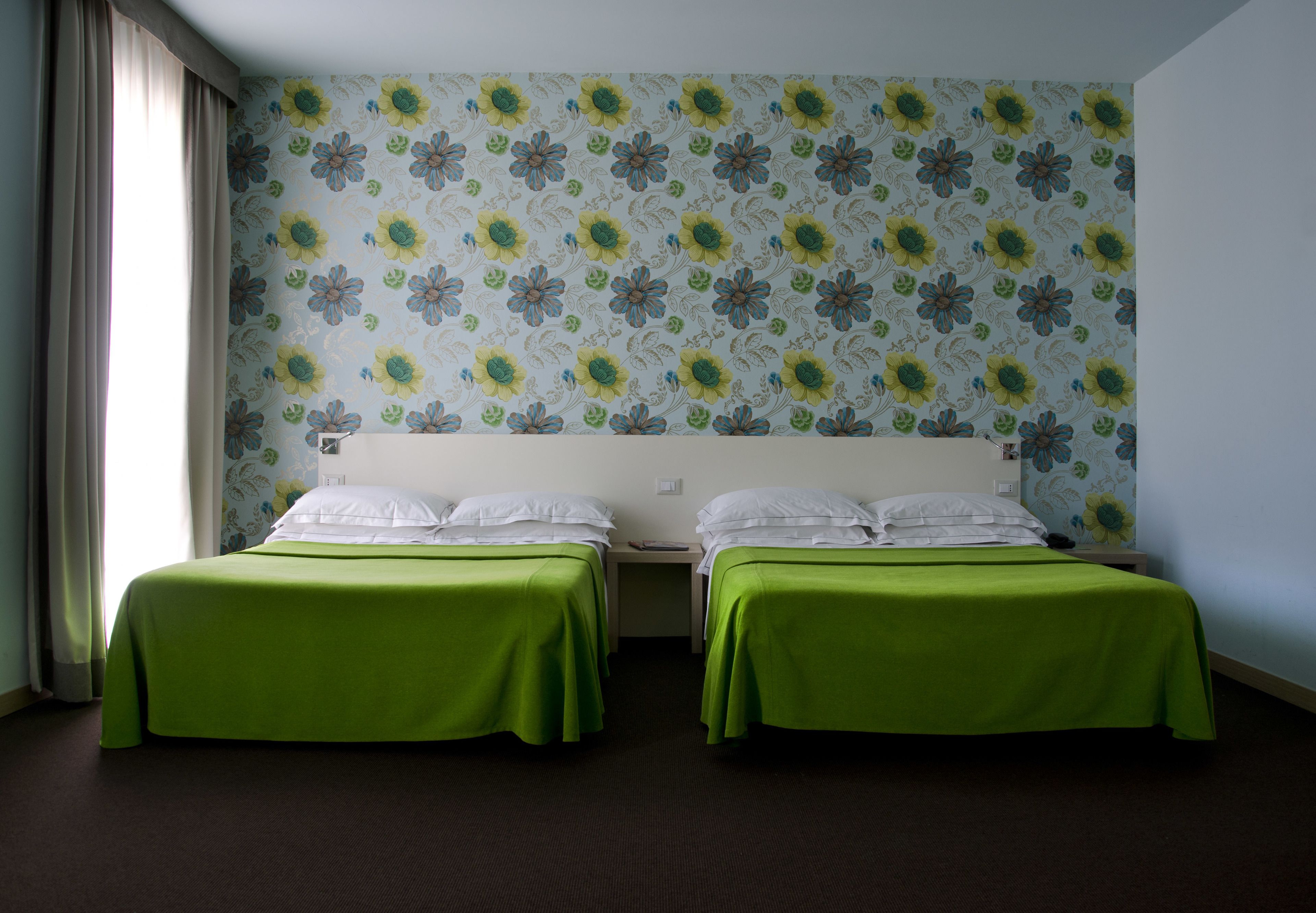 A hotel room with two single beds, a floral wallpaper, and a window with curtains.