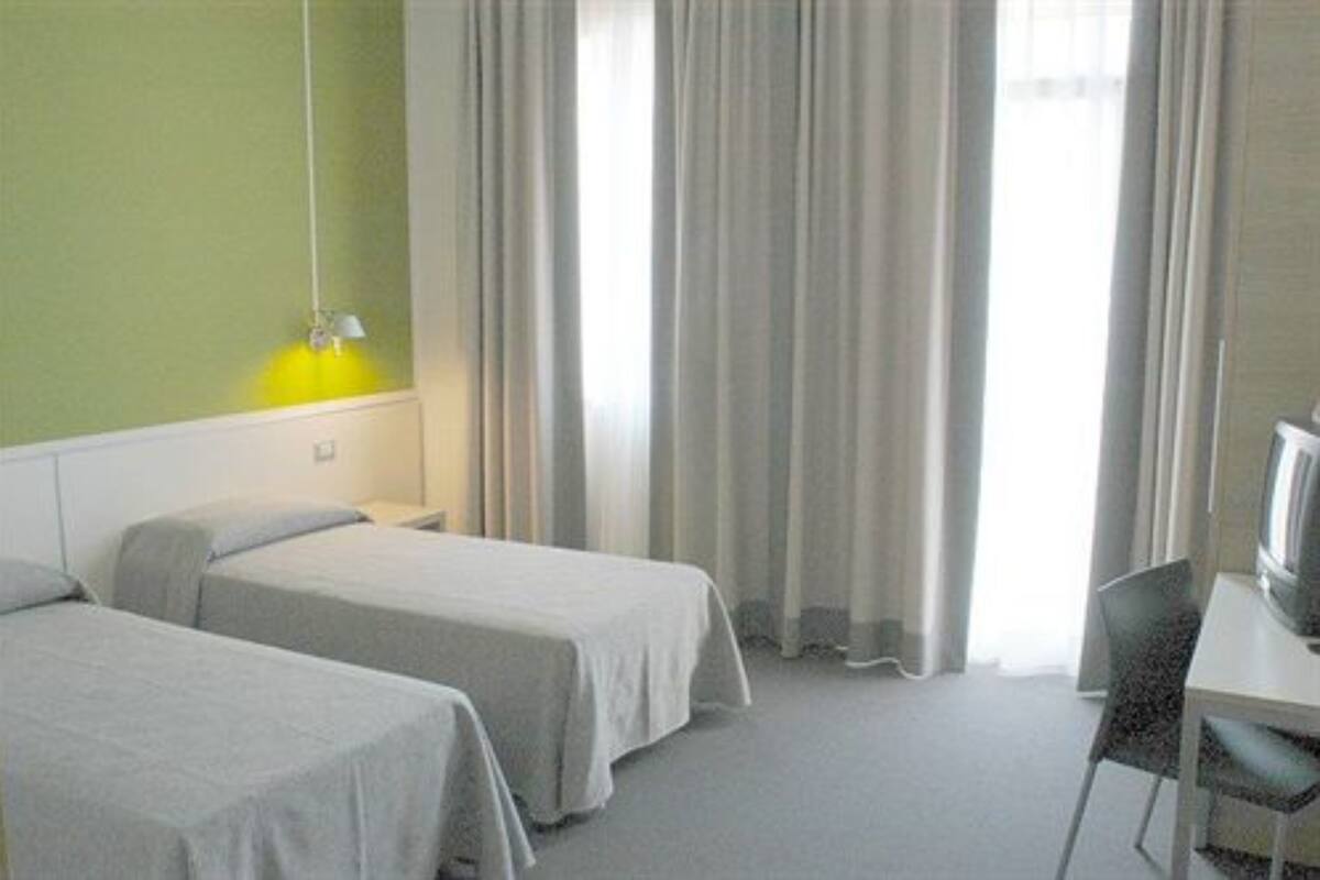 Una camera d'albergo con due letti singoli, una scrivania con una sedia, una televisione e una finestra con tende.