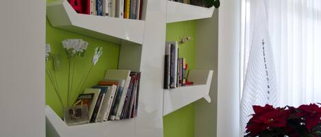 Un salón con una pared verde de acento, estantes blancos flotantes con libros y objetos decorativos, un sofá negro con un cojín estampado y una ventana con cortinas translúcidas.