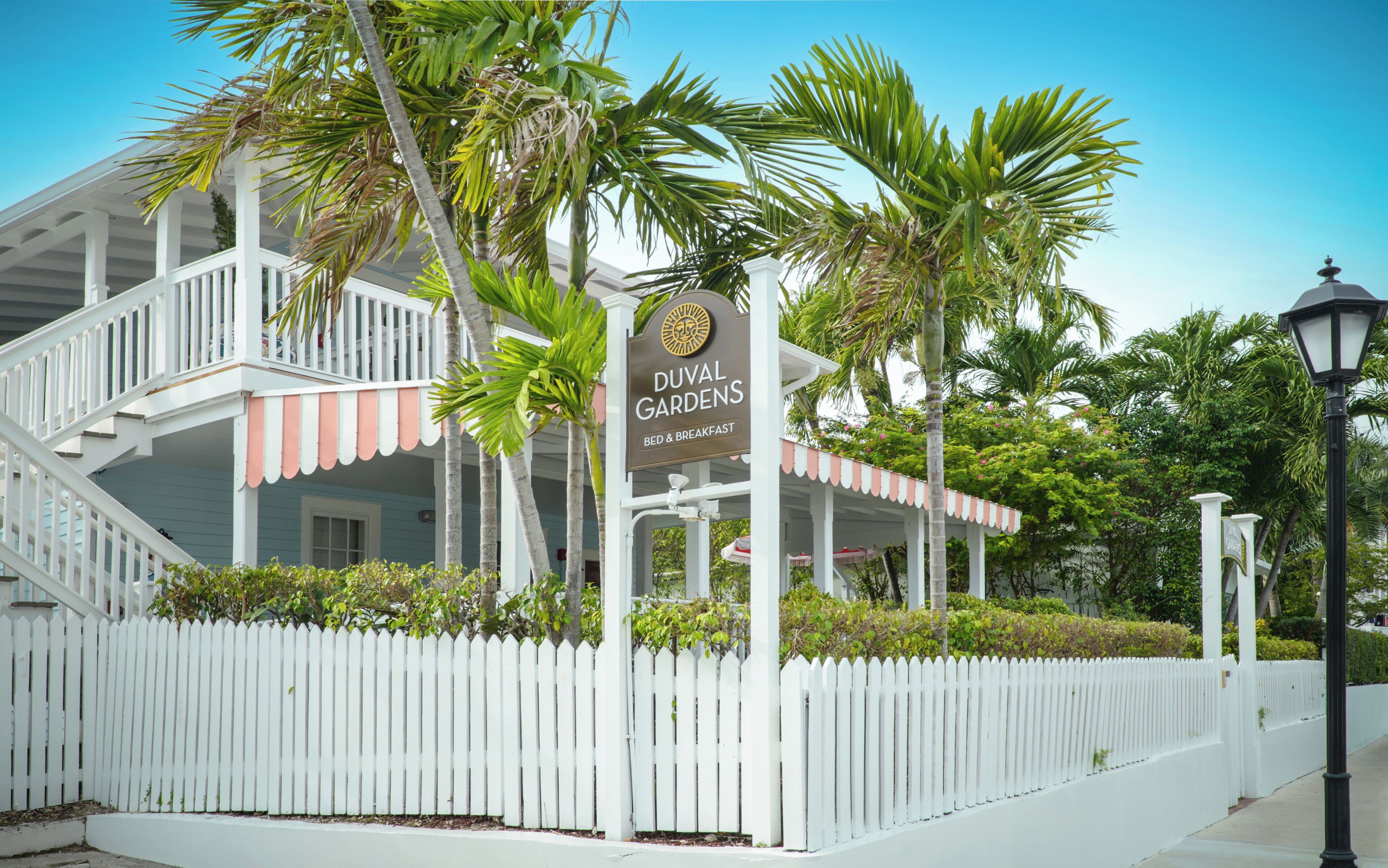 Une clôture blanche en bois, un panneau indiquant « Duval Gardens Bed & Breakfast », et un palmier.