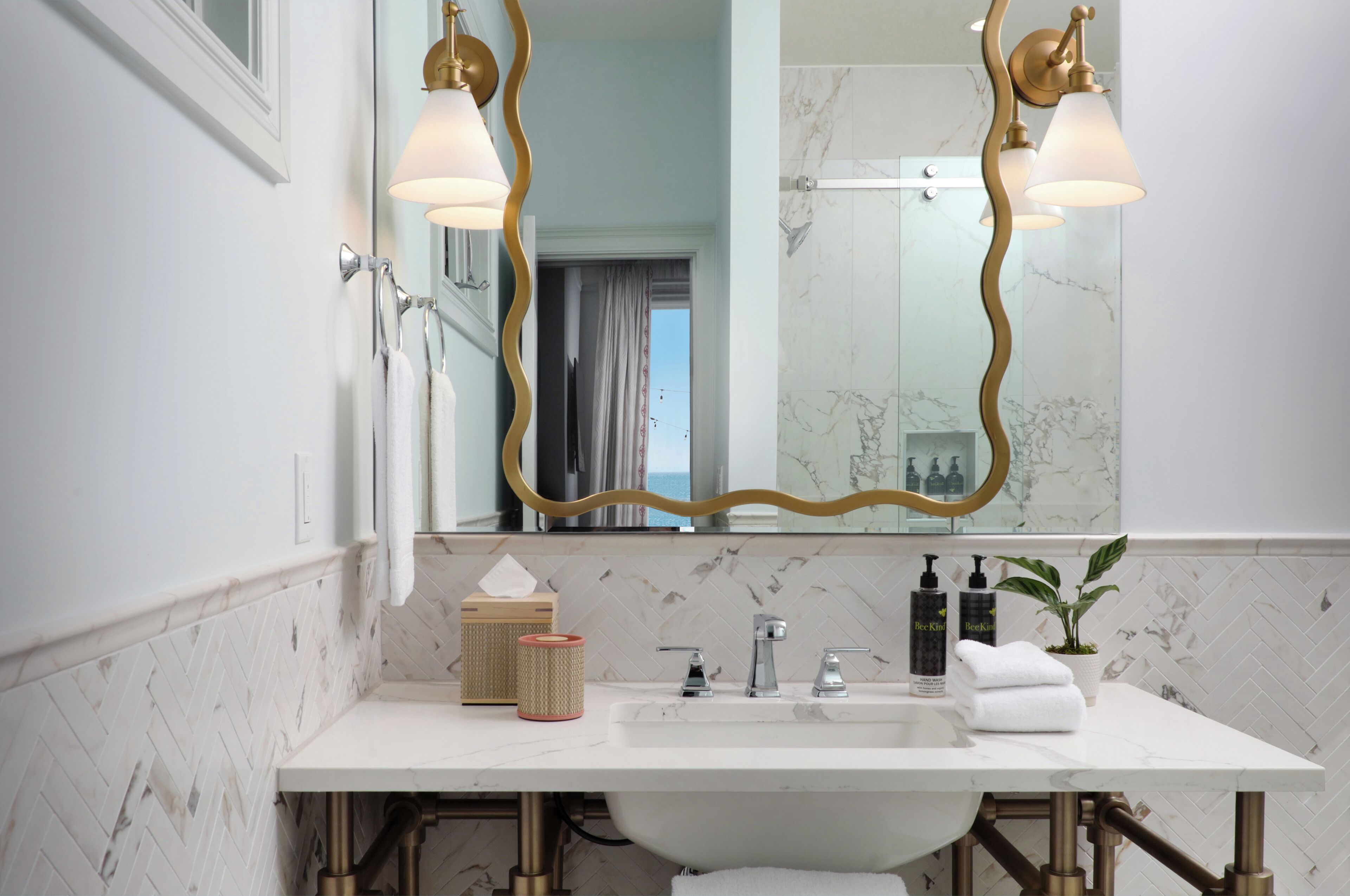 Une salle de bain avec un lavabo blanc, deux robinets, un miroir avec un cadre doré et un porte-serviettes.