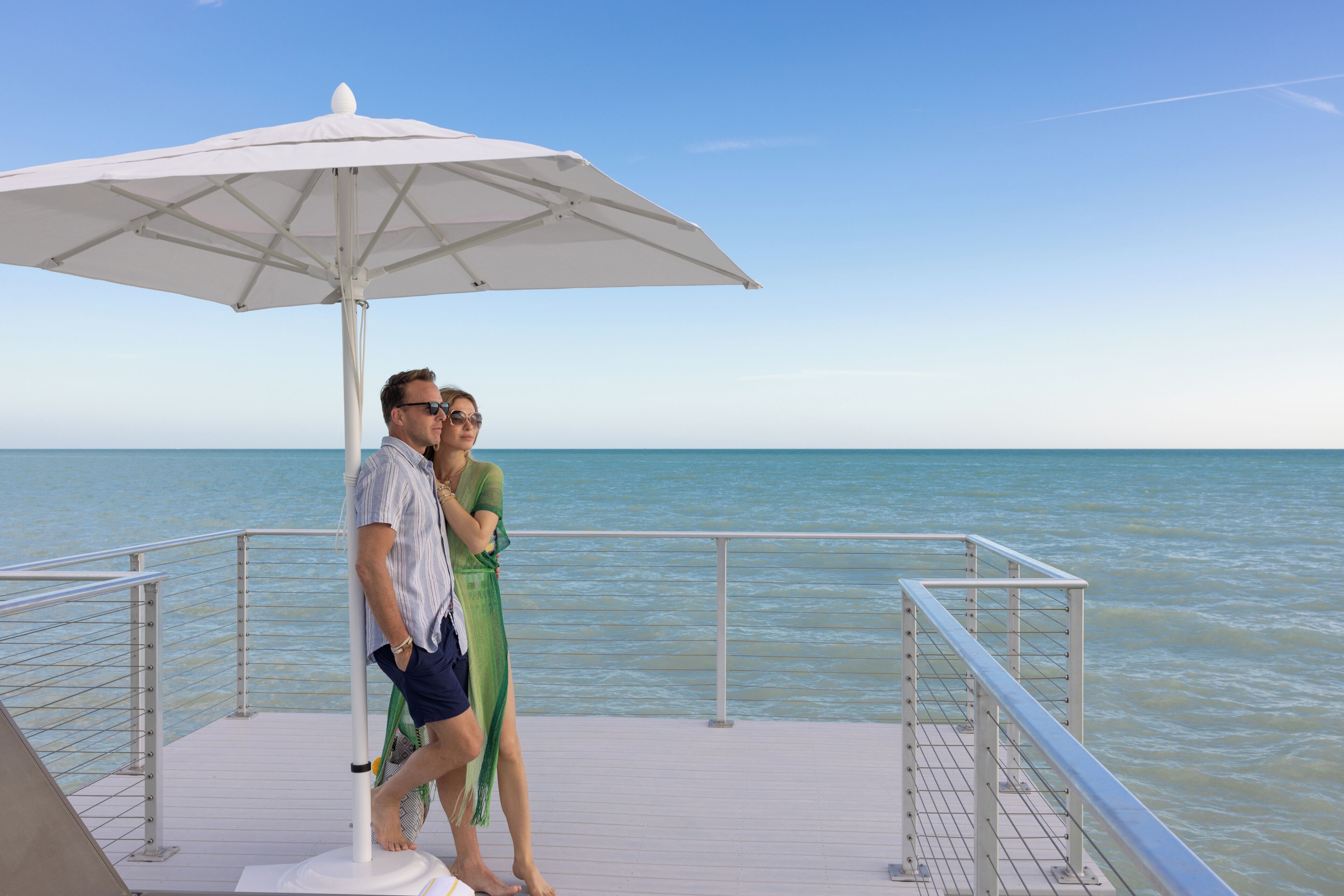 Ein Paar steht unter einem weißen Sonnenschirm auf einem Bootsdeck mit Geländer und blickt auf das Meer.