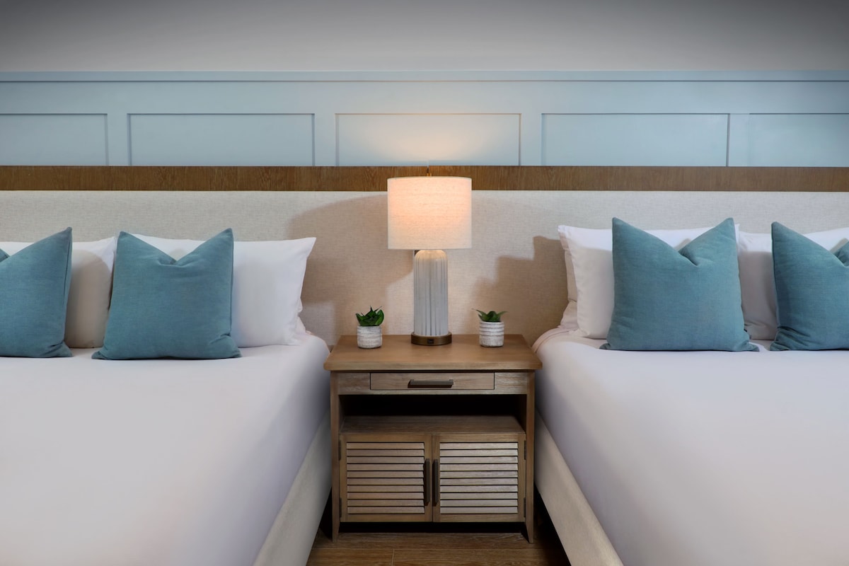 Une chambre d’hôtel avec deux lits, une table de chevet avec une lampe et une tête de lit ornée d’un panneau en bois.