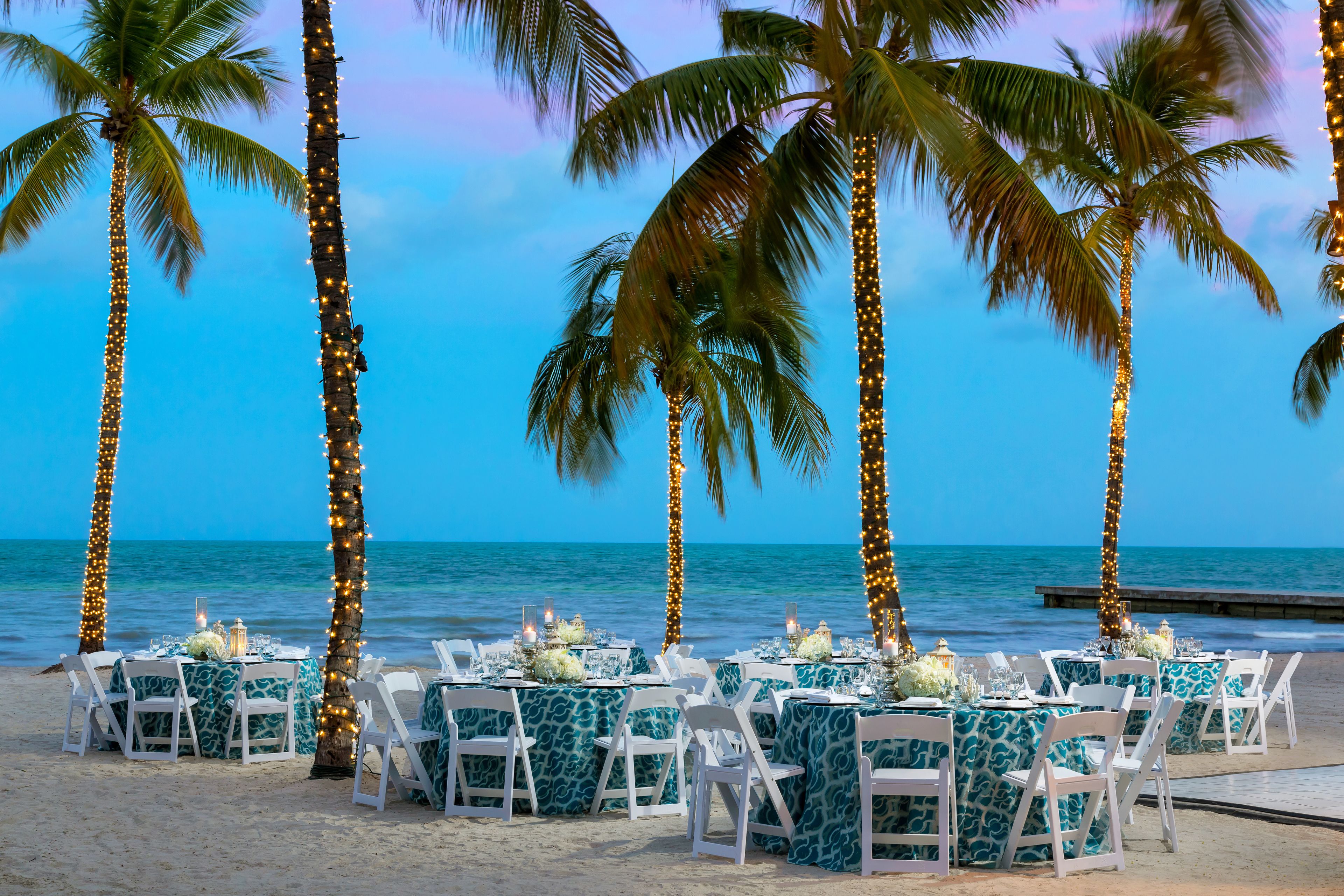 A beachside dining setup with tables and chairs, palm trees, and a view of the ocean.