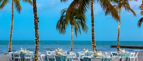 A beachside dining setup with tables and chairs, palm trees, and a view of the ocean.