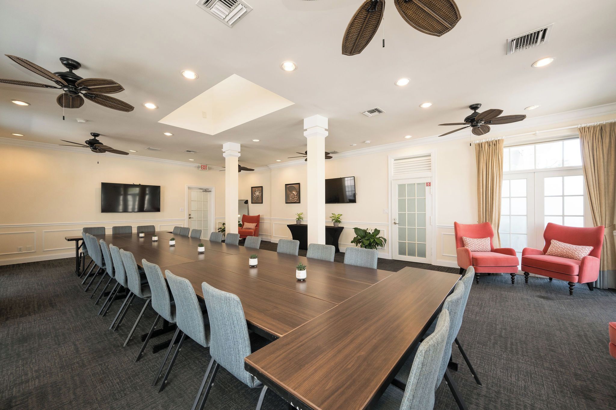 A conference room with a long table, chairs, ceiling fans, recessed lighting, and a large window with curtains.