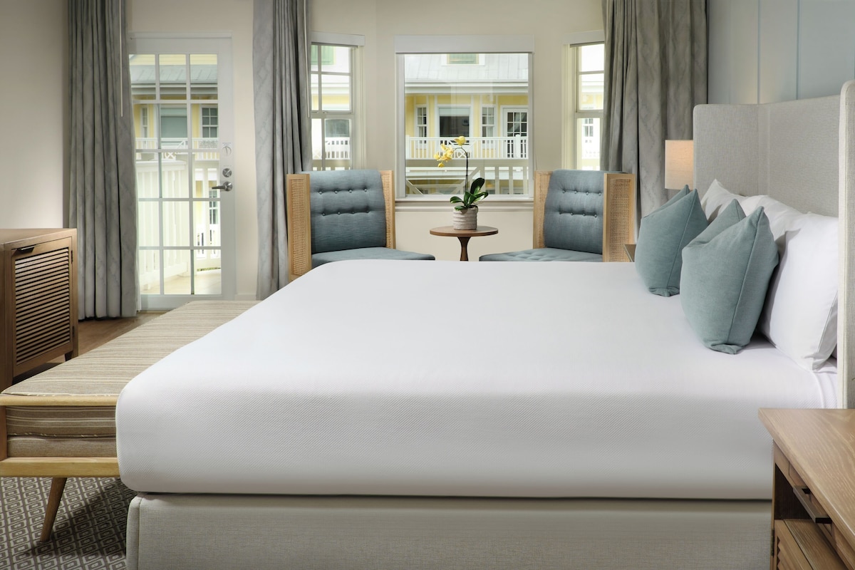 Une chambre d’hôtel avec un grand lit, deux fauteuils, une petite table et une vue sur un immeuble par la fenêtre.
