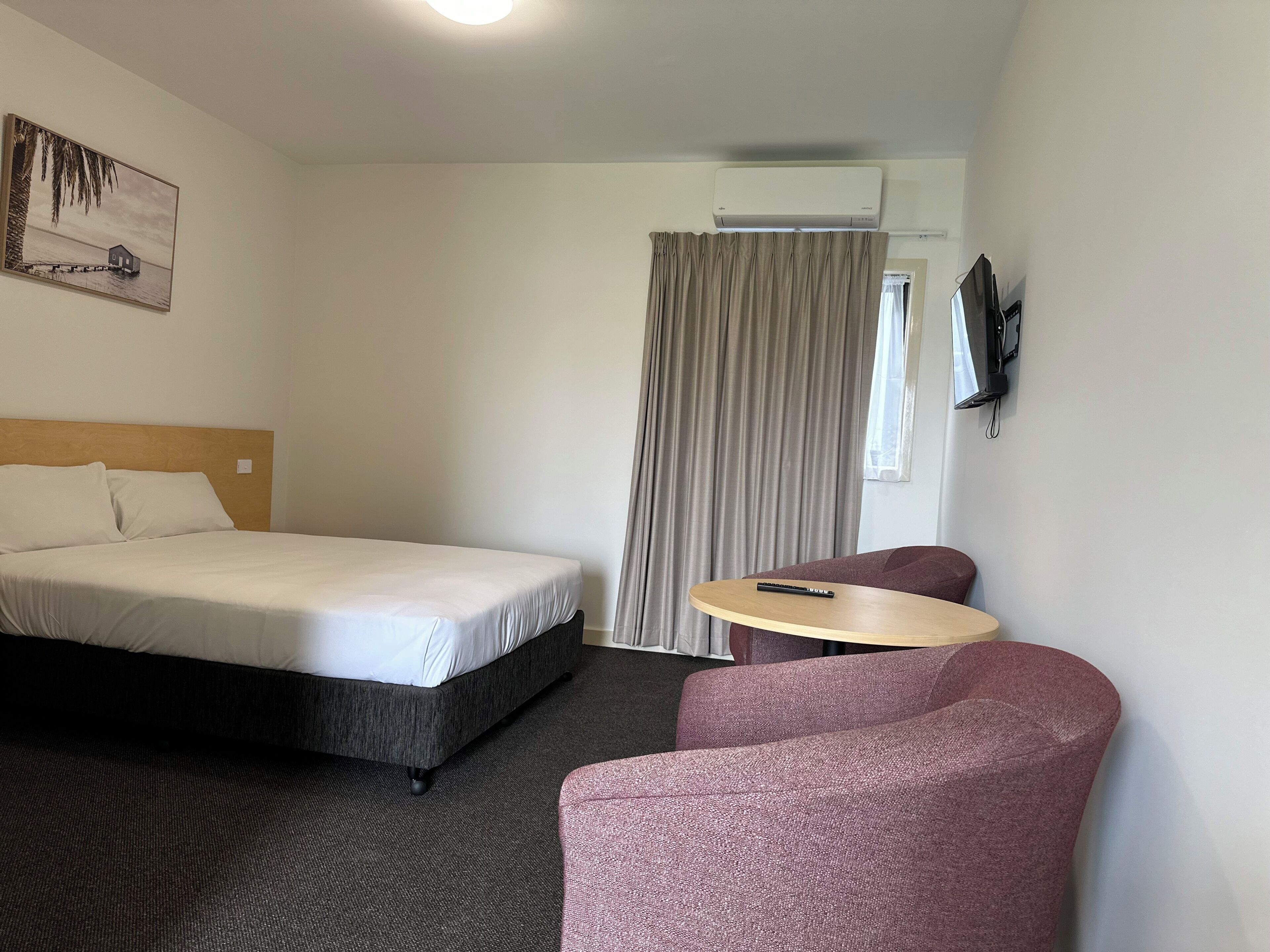 A hotel room with a bed, a small table, a chair, an air conditioning unit, and a painting on the wall.