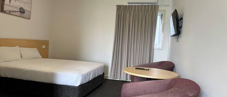 A hotel room with a bed, a small table, a chair, an air conditioning unit, and a painting on the wall.