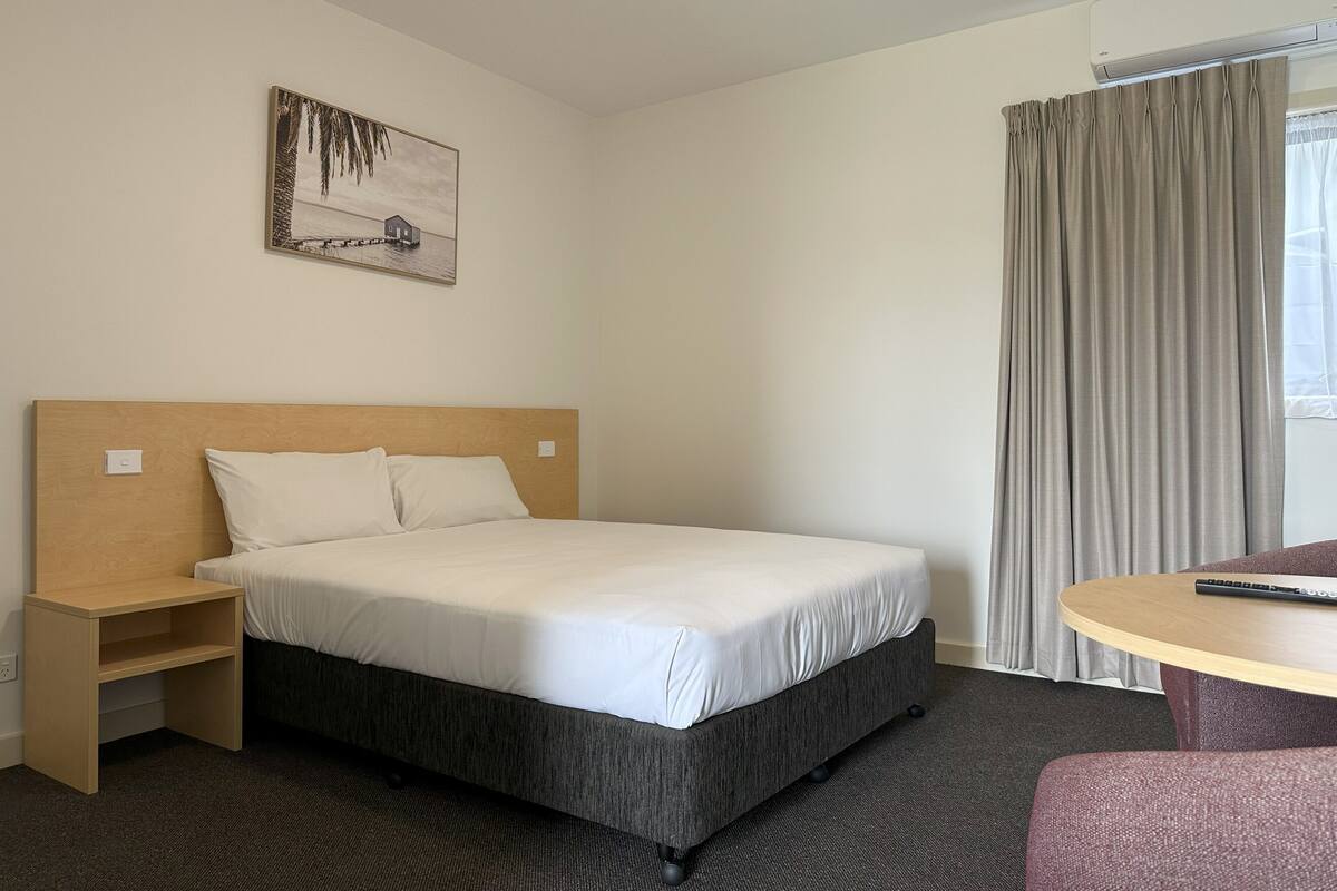 A hotel room with a single bed, a wooden nightstand, a small table, and a chair.
