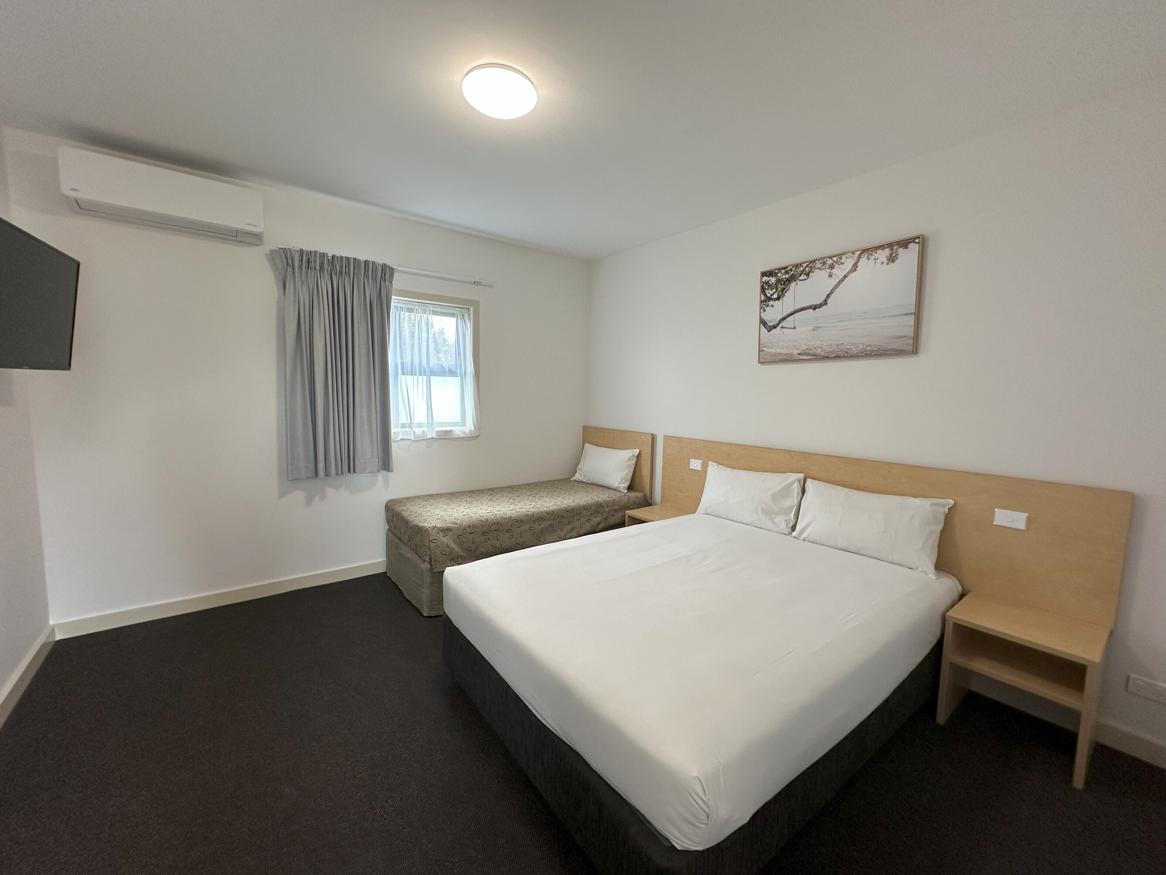 A hotel room with a large bed, a smaller bed, a bedside table, a nightstand, a window with curtains, an air conditioning unit, and a wall-mounted picture.