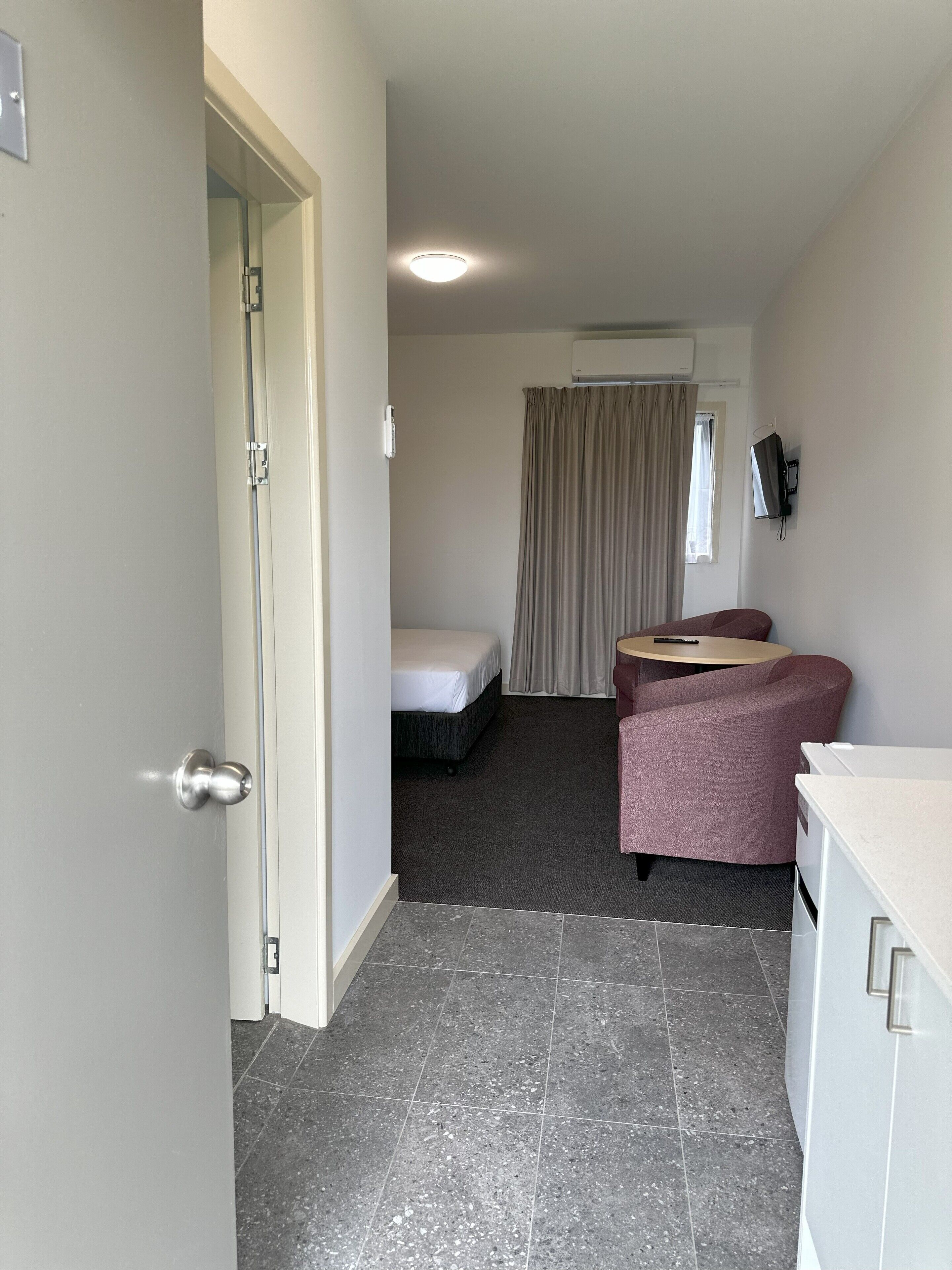A compact hotel room with a bed, a small seating area, a kitchenette, and a bathroom.