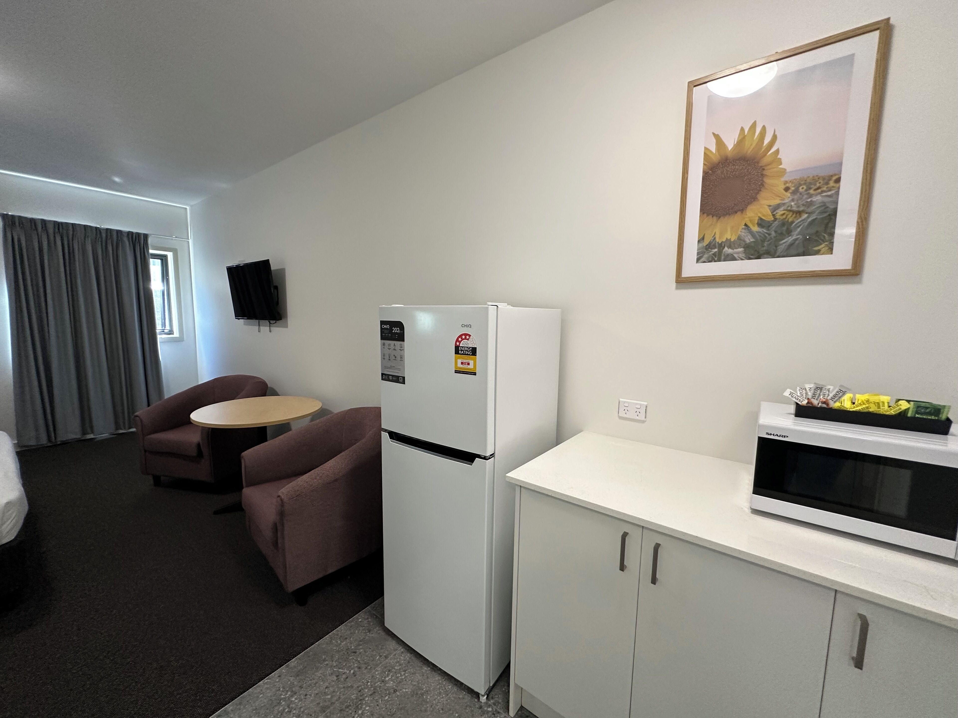Habitación de hotel compacta con kitchenette, zona de comedor con una mesa y dos sillas, refrigerador, microondas y televisor montado en la pared.