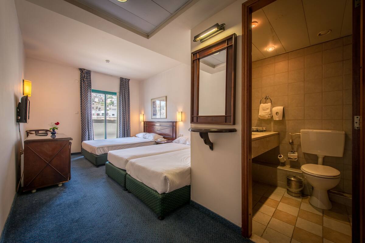 A hotel room with two beds, a desk with a lamp, a mirror, and a bathroom with a toilet and sink.