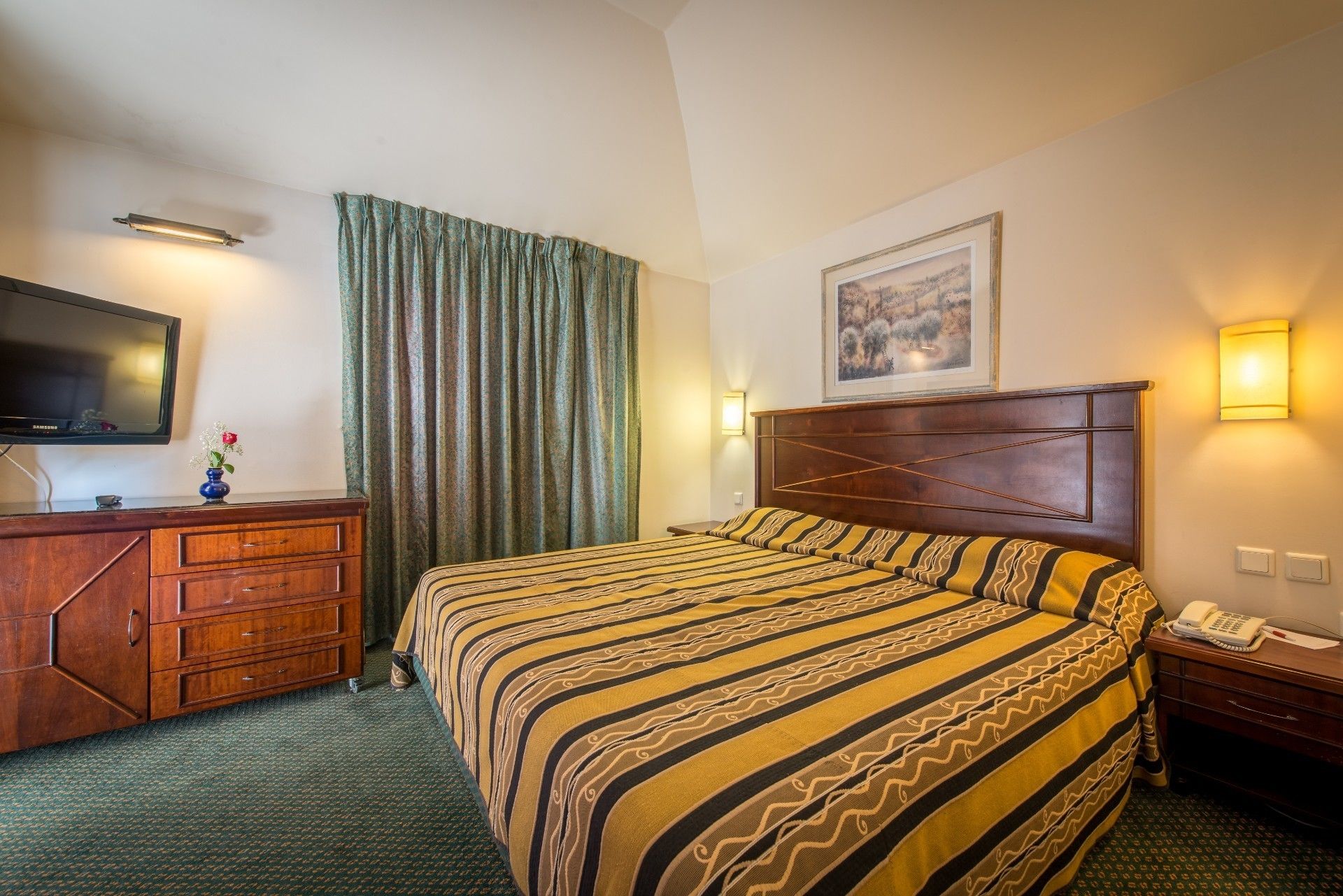A hotel room with a bed, a TV, a wooden dresser, a telephone, and a painting on the wall.