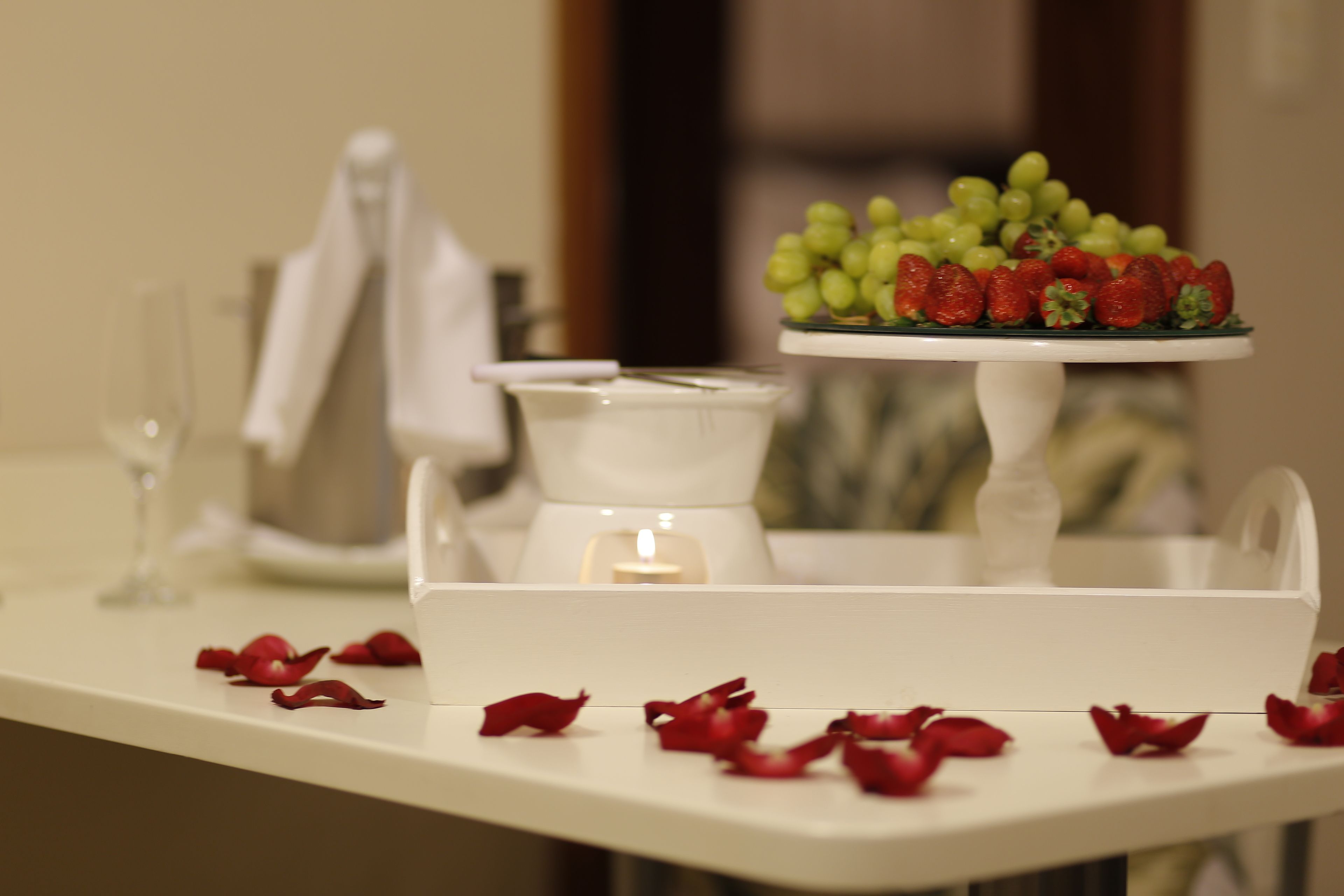 Uma mesa de sobremesas com um suporte branco para bolo, uma vela e pétalas de rosa espalhadas.