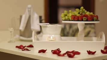 Uma mesa de sobremesas com um suporte branco para bolo, uma vela e pétalas de rosa espalhadas.