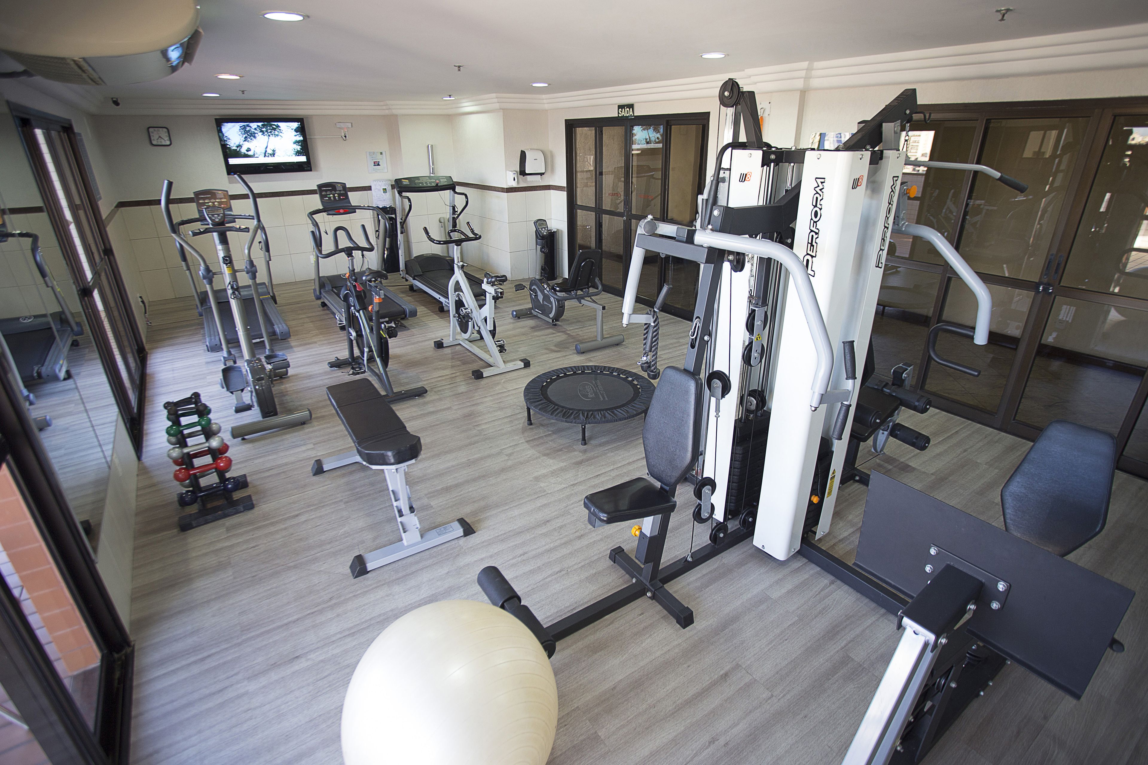 Un gimnasio con diferentes máquinas de ejercicio, una pelota grande de entrenamiento y un televisor en la pared.