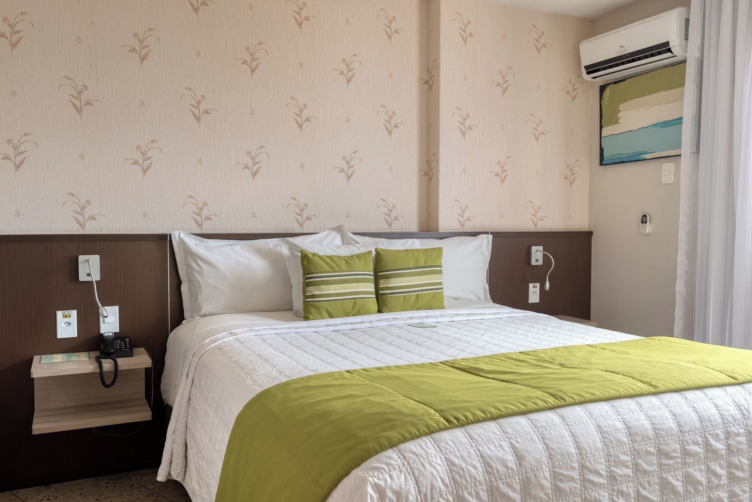 Habitación de hotel con cama, mesita de noche con teléfono, enchufe y aire acondicionado.