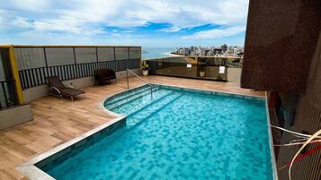 Piscina en la azotea con vistas a la ciudad, terraza de madera y sillones de descanso.