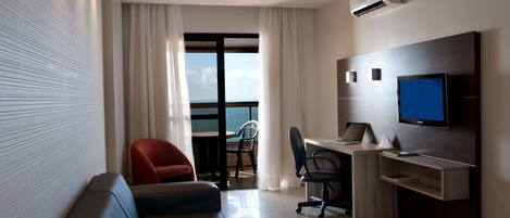 A modern hotel room with a sofa, desk, chair, TV, and a view of the sea through a large window.