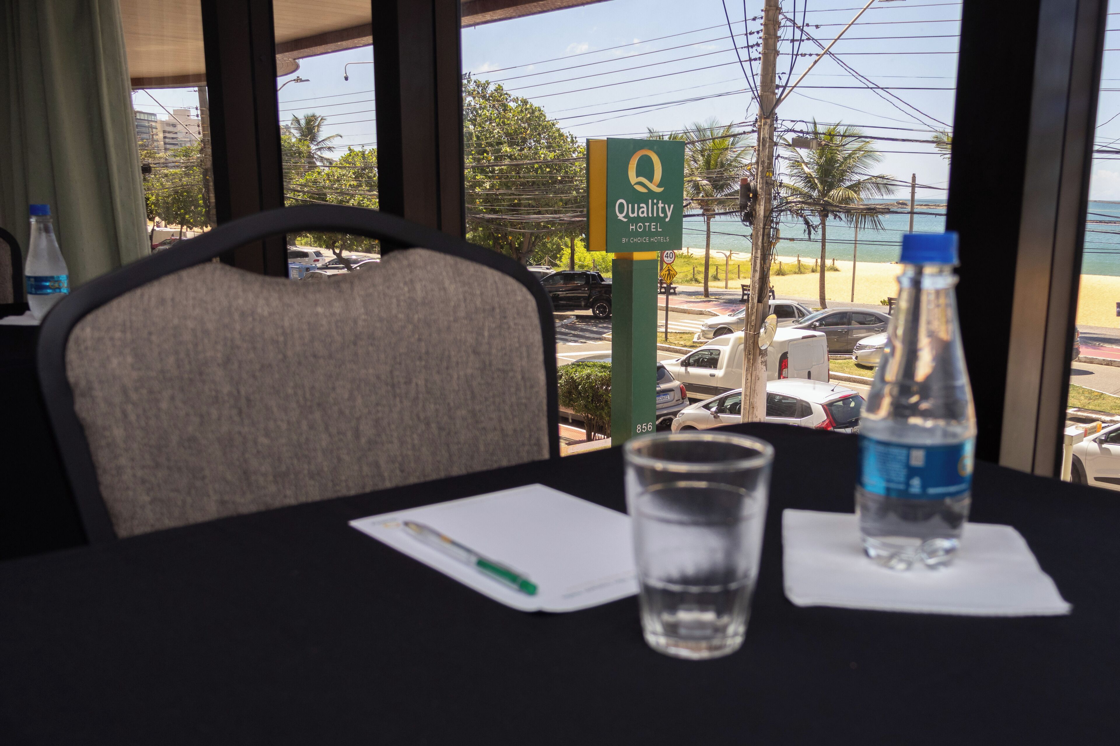 Quarto de hotel com vista para a praia, uma mesa com um copo de água, um bloco de notas e uma caneta.