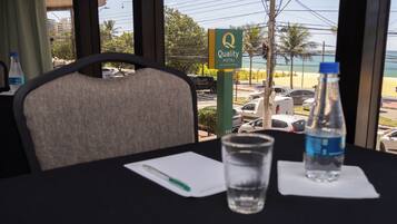 Quarto de hotel com vista para a praia, uma mesa com um copo de água, um bloco de notas e uma caneta.