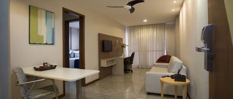 Una habitación de hotel moderna con zona de comedor, sofá, televisión montada en la pared y ventilador de techo.