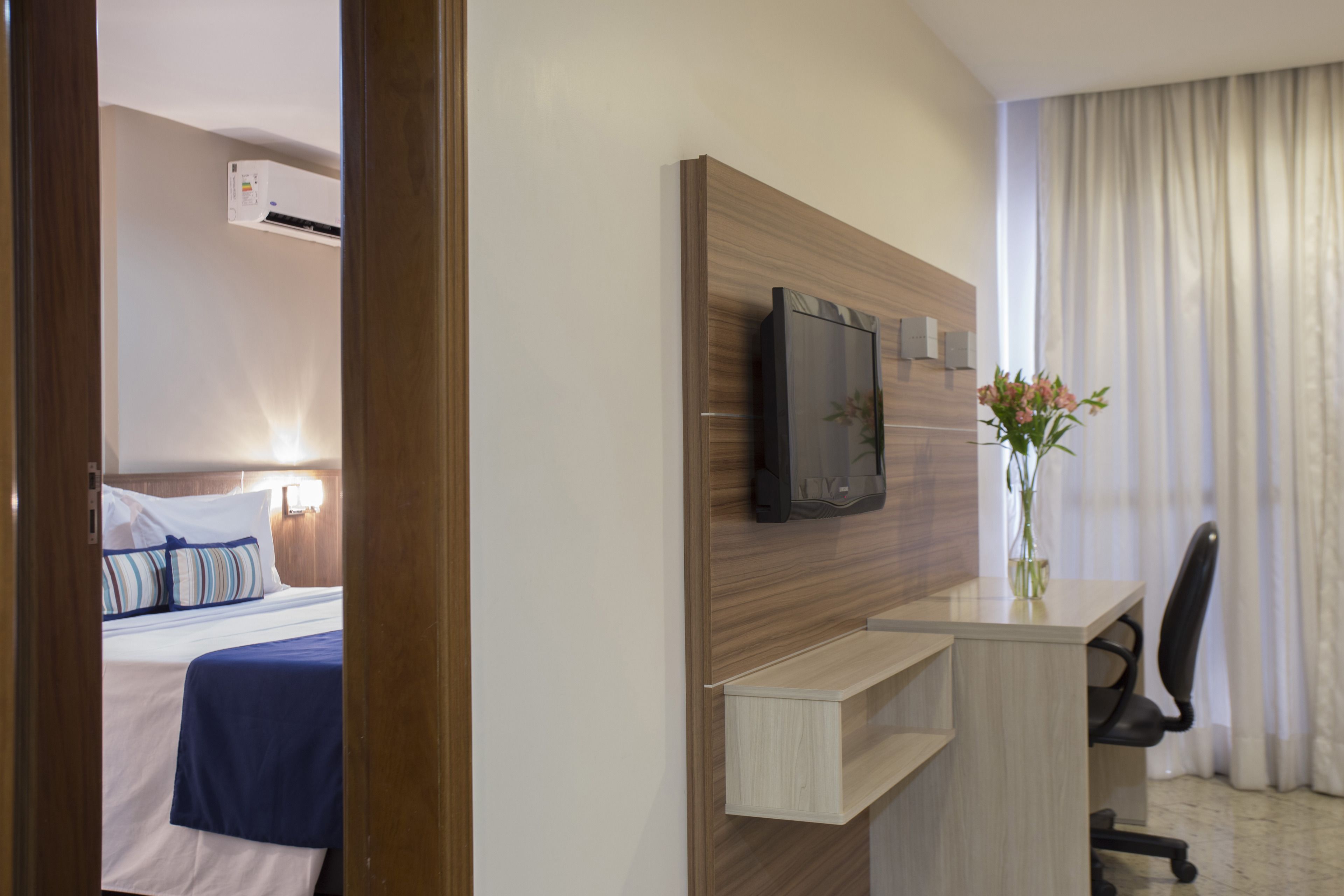 Una habitación de hotel con cama, escritorio con silla, televisor y un florero con flores.