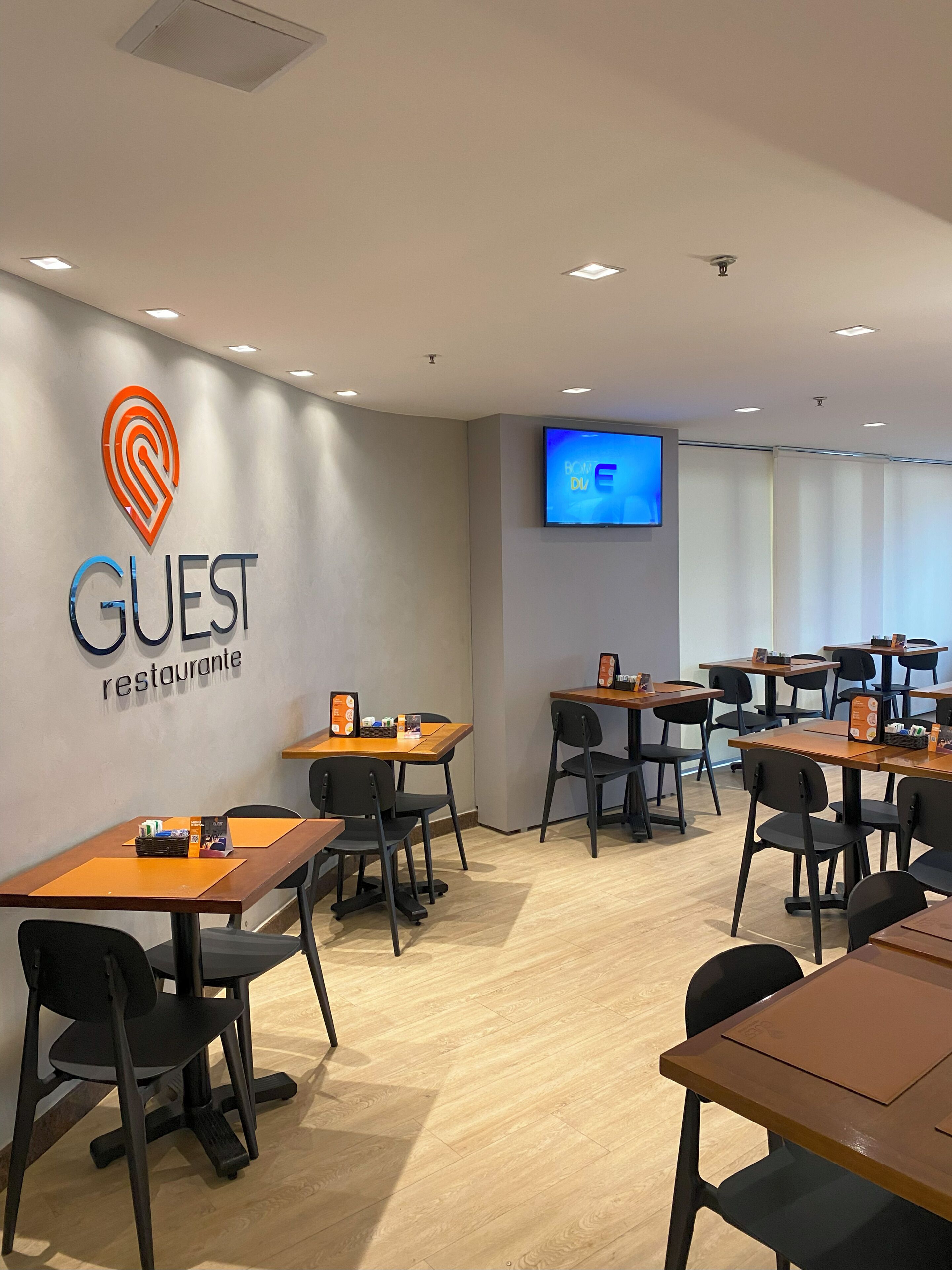 Ein modernes Restaurant mit Tischen, Stühlen und einem Logo an der Wand, sowie einem Fernsehbildschirm.